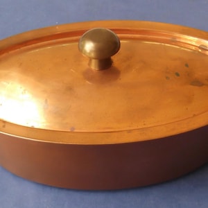 May include: An oval copper cooking pot with a matching lid and two side handles. The pot features a polished metallic finish with a warm reddish-brown hue. A rounded knob is centered on the lid, and curved handles are attached to each side.