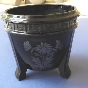 May include: A black planter with a decorative Greek key border and three legs. The planter features a floral design etched on the front. The planter is made of a glossy material.