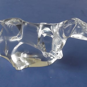 May include: A clear glass sculpture of a dog in a reclining position. The dog's form is stylized, with smooth, flowing lines. The sculpture is transparent, allowing light to pass through, and sits on a blue surface.