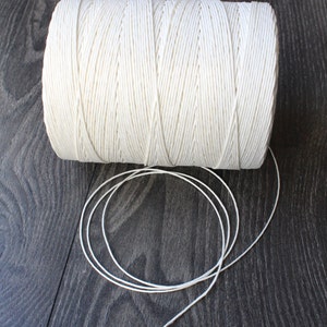 Może przedstawiać: Szpula białego sznurka bawełnianego, z kawałkiem sznurka rozwiniętym i leżącym na ciemnym drewnianym podłożu.