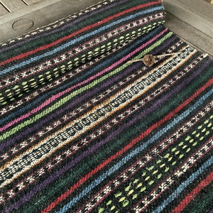 May include: A rolled-up, handwoven textile with a striped pattern. The design features alternating bands of black, red, blue, green, purple, and white. The textile has a textured appearance and a small pinecone rests on top.