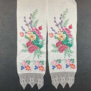 May include: A pair of white rectangular fabric panels featuring a cross-stitched floral bouquet design in shades of pink, red, yellow, purple, and green. The bottom edges are finished with a decorative white crocheted lace trim with points.