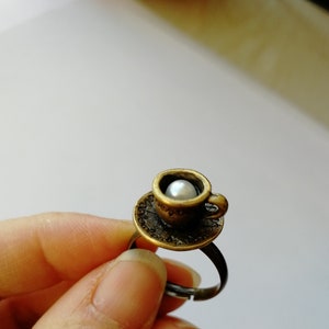 Könnte beinhalten: Ein bronzefarbener Ring mit einem Miniatur-Tassen-Design. Die Tasse, mit einer Perle darin, sitzt auf einer Untertasse. Der Ring ist verstellbar. Tasse und Untertasse weisen aufwendige Details auf, die eine Vintage-Ästhetik erzeugen.