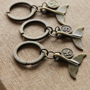 May include: Three bronze-colored keychains with a whale tail design. Each keychain has a circular ring and a small circular charm with a letter. The letters are "N", "S", and "K".
