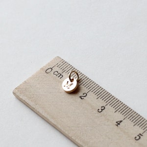 Op de afbeelding: Een kleine, goudkleurige bedel met de letter "M" gegraveerd op een ronde hanger. De bedel is bevestigd aan een kleine lus. Een houten liniaal met centimeter markeringen staat op de achtergrond.