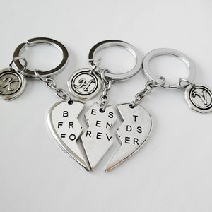 Op de afbeelding: Drie zilverkleurige sleutelhangers met een gebroken hart ontwerp met de tekst "Best Friends Forever". Elke sleutelhanger heeft een cirkelvormige bedel met een letter: "K", "H" en "N". De sleutelhangers zijn verbonden door kleine kettingen.
