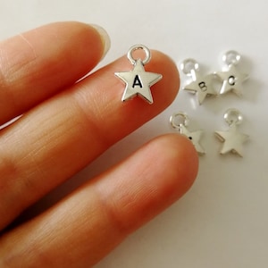 May include: Small silver star charms, each with a loop for hanging. One charm features the letter "A", while others display "B" and "C". The charms are metallic and suitable for jewellery or craft projects.