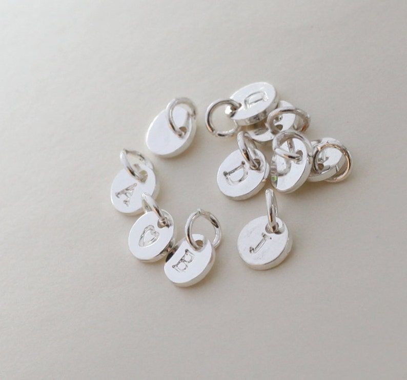 Op de afbeelding: Een verzameling kleine, zilverkleurige initiaalbedels. Elke bedel is een kleine, ronde schijf met een enkele letter gegraveerd. De bedels hebben een lus aan de bovenkant om aan een ketting of armband te bevestigen. De achtergrond is lichtbeige.
