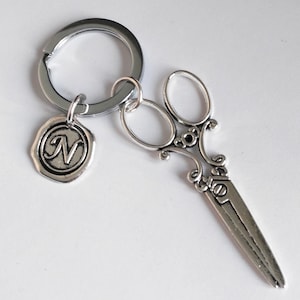 May include: Silver-toned keychain featuring a pair of decorative scissors charm and a circular charm with the letter "N". The scissors have ornate handles and a detailed design. The keychain ring is also silver.