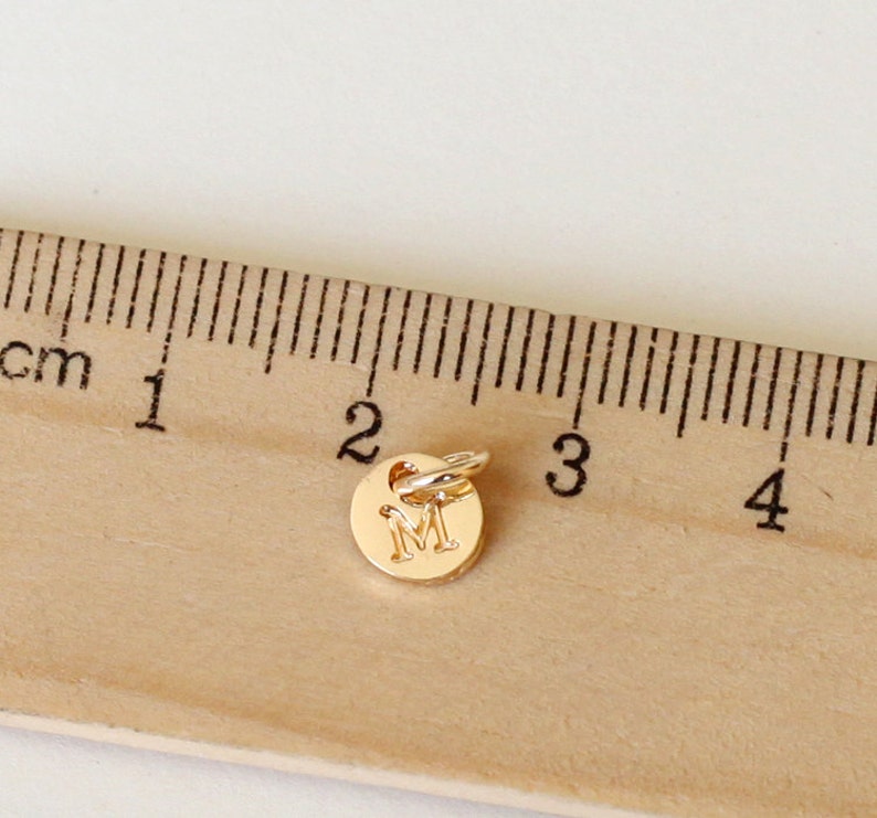 May include: A small, gold-colored charm with the letter "M" engraved on a circular pendant. The charm is approximately 0.4 inches in diameter and has a small loop for attaching to a necklace or bracelet. The charm is shown next to a ruler.