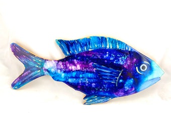 May include: A blue and purple fish painted on wood with gold accents. The fish has a white eye and is painted in a watercolor style.