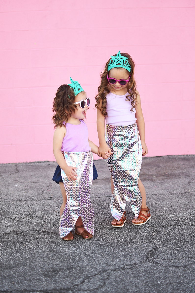 May include: Two young girls wearing mermaid tails and matching green crowns with star accents. They are both wearing sunglasses and smiling. The girls are standing on a pink background.