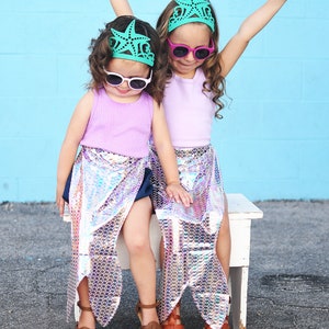 May include: Two young girls wearing mermaid tails and teal starfish crowns, with pink and green sunglasses, sitting on a white bench.