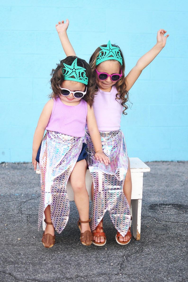 May include: Two young girls wearing mermaid tails and teal starfish crowns, with pink and green sunglasses, sitting on a white bench.