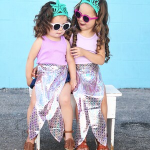 May include: Two girls wearing mermaid tails and matching starfish crowns, one with pink sunglasses and the other with white sunglasses, sitting on a white bench.