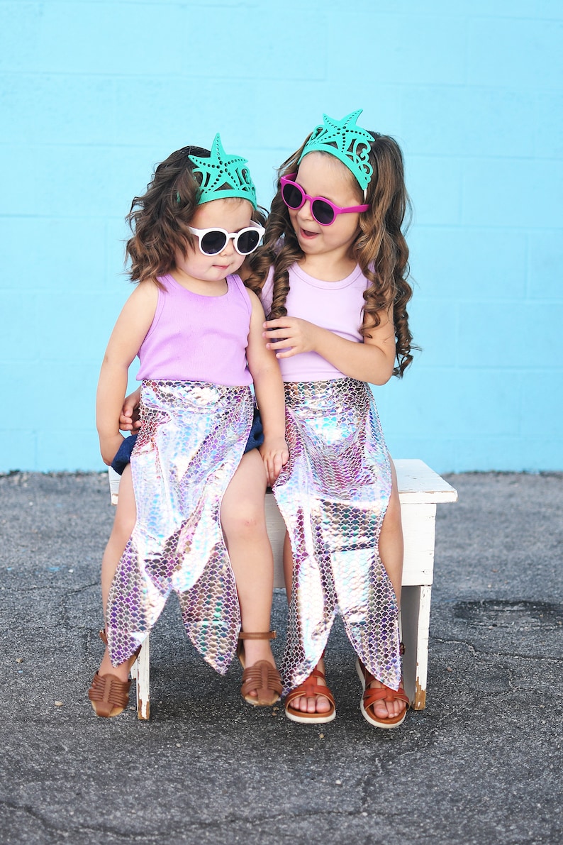 May include: Two girls wearing mermaid tails and matching starfish crowns, one with pink sunglasses and the other with white sunglasses, sitting on a white bench.