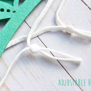 May include: Close-up of a white adjustable headband with white plastic sliders. The headband is made of elastic material and is designed to fit comfortably around the head. "ADJUSTABLE HEADBAND" is written in green text below the headband.