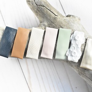 May include: A collection of rectangular hair clips in various colors, including navy blue, tan, cream, pale pink, light green, and a white lace design. The clips are arranged on a piece of driftwood against a white background.