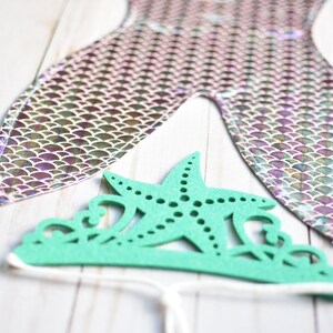 May include: A green felt mermaid crown with a starfish design. The crown is tied with a white string.