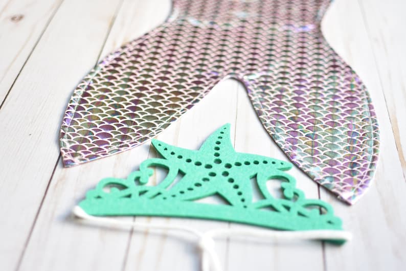 May include: A green felt mermaid crown with a starfish design. The crown is tied with a white string.