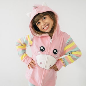 May include: A pink unicorn hoodie with a white face and large eyes. The hoodie has a hood with white ears and a zipper closure. The sleeves are striped with pastel colors. The child is smiling and wearing the hoodie.