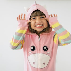 May include: A pink unicorn onesie with a hood, ears, and a horn. The sleeves have pastel rainbow stripes. The onesie features a unicorn face on the front with large eyes and a heart. A child is wearing the onesie and making a silly face.