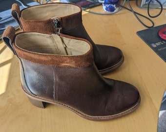 May include: A pair of brown leather ankle boots with a suede trim around the top. The boots have a zipper on the inside of each boot and a block heel.