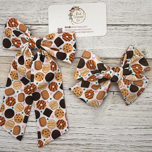 May include: Two fabric hair bows with a cookie pattern. The bows feature various cookie designs in brown, tan, and white, with small pink hearts. One bow is larger with long tails, and the other is smaller. A business card is in the background.