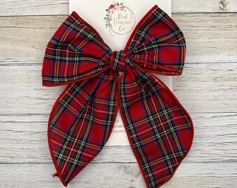May include: A large, red plaid bow with a classic pattern of green, blue, and yellow lines. The bow is centered on a white card with the text "Red Treasure Co." and a floral design. The bow is made of fabric.