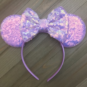Puede incluir: Una diadema de lentejuelas lavanda con orejas de ratón y un lazo. Las orejas y el lazo están cubiertos de lentejuelas iridiscentes, creando un efecto brillante. La diadema es de color lavanda liso.
