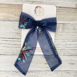 May include: A navy blue mesh hair bow featuring embroidered firework-like patterns in red, silver, and light blue sequins. The bow is attached to a white display card with the text Red Treasure Co and social media handles printed on it.