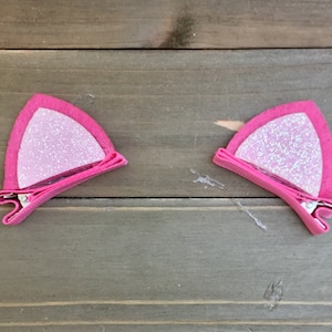 May include: Two pink felt cat ears with glitter accents, each attached to a metal clip for hair.