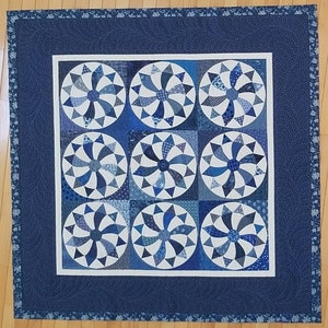Könnte beinhalten: Eine blaue und weiße Steppdecke mit einem sich wiederholenden Muster aus Windrädern. Die Steppdecke ist mit einem dunkelblauen Rand eingefasst.