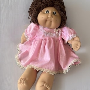 Op de afbeelding: Een vintage pop met bruin haar en een rond gezicht. De pop draagt een roze jurk met witte stippen en kanten afwerking. De armen en benen van de pop zijn gemaakt van een zachte, beige stof.