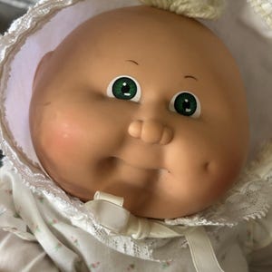 Puede incluir: Una muñeca bebé vintage con cara redonda, mejillas rosadas y ojos verdes. La muñeca lleva un gorro blanco con ribete de encaje y un vestido con estampado floral. Se ve un pequeño mechón de pelo rubio.