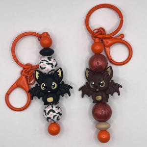 May include: Two Halloween-themed keychains. One features a black bat charm, the other a brown bat. Both have orange hardware, decorative beads, and a lobster clasp. The beads include black, white, and orange colors.