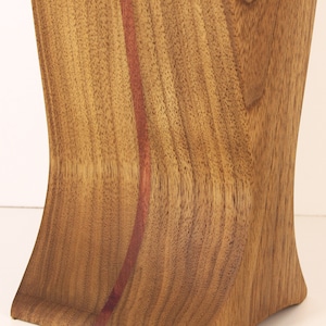 K&ouml;nnte beinhalten: Eine Holzvase mit einer geschwungenen Form und einer rotbraunen Holzmaserung. Die Vase hat eine d&uuml;nne, vertikale Linie aus dunklerem Holz, die in der Mitte verl&auml;uft.