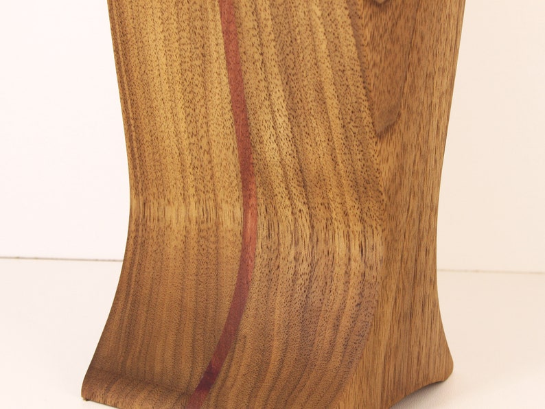 K&ouml;nnte beinhalten: Eine Holzvase mit einer geschwungenen Form und einer rotbraunen Holzmaserung. Die Vase hat eine d&uuml;nne, vertikale Linie aus dunklerem Holz, die in der Mitte verl&auml;uft.