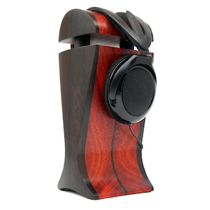Puede incluir: Un soporte para auriculares de madera con un patrón de veta de madera negro y rojo. El soporte sostiene un par de auriculares negros con una copa de malla.