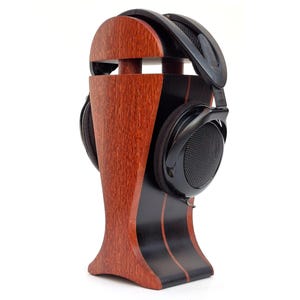 K&ouml;nnte beinhalten: Ein Kopfh&ouml;rerst&auml;nder aus Holz mit dunklem Finish und einem Paar schwarzer Kopfh&ouml;rer. Der St&auml;nder besteht aus rotbraunem Holz mit schwarzen Akzenten und einem geschwungenen Design. Die Kopfh&ouml;rer liegen auf dem St&auml;nder.