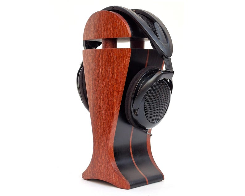 K&ouml;nnte beinhalten: Ein Kopfh&ouml;rerst&auml;nder aus Holz mit dunklem Finish und einem Paar schwarzer Kopfh&ouml;rer. Der St&auml;nder besteht aus rotbraunem Holz mit schwarzen Akzenten und einem geschwungenen Design. Die Kopfh&ouml;rer liegen auf dem St&auml;nder.