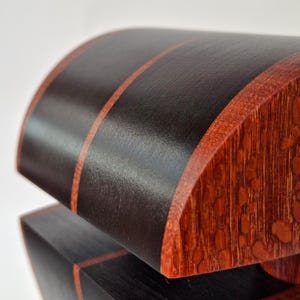 K&ouml;nnte beinhalten: Nahaufnahme einer handgefertigten Holzkiste mit gew&ouml;lbtem Deckel. Die Kiste hat eine dunkelbraune Oberfl&auml;che mit zwei d&uuml;nnen roten Streifen auf dem Deckel und rot get&ouml;nten Holzakzenten an den Kanten. Die Kiste hat ein geometrisches Design.