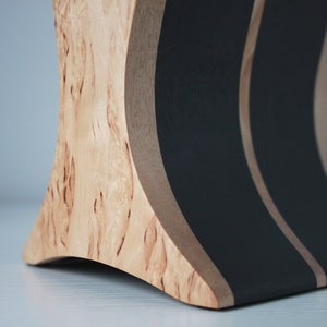 May include: A wooden stand with a curved design, featuring black painted stripes. The stand is made of light wood with a natural grain pattern.