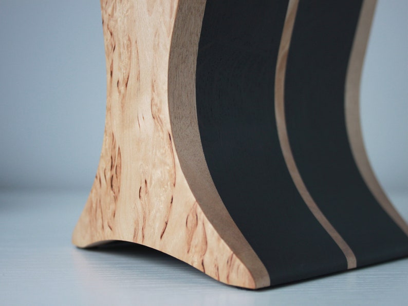 May include: A wooden stand with a curved design, featuring black painted stripes. The stand is made of light wood with a natural grain pattern.