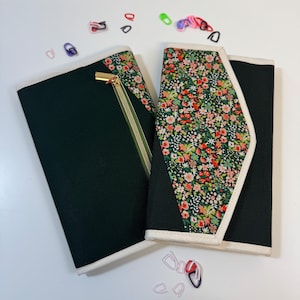 Pode incluir: Dois organizadores de tecido retangulares com pain&eacute;is pretos e um padr&atilde;o floral com pequenas flores vermelhas, rosas e brancas sobre um fundo verde. Um organizador tem um fecho dourado e ambos s&atilde;o rematados com um debrum branco. Pequenos marcadores de pontos de pl&aacute;stico coloridos est&atilde;o espalhados &agrave; volta dos objetos.