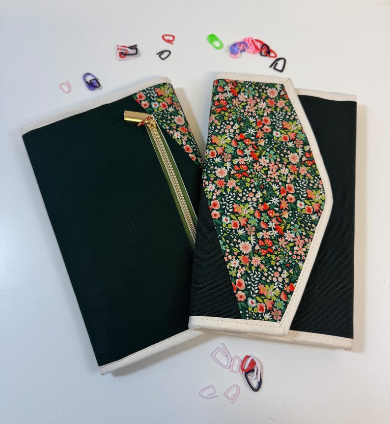 Pode incluir: Dois organizadores de tecido retangulares com pain&eacute;is pretos e um padr&atilde;o floral com pequenas flores vermelhas, rosas e brancas sobre um fundo verde. Um organizador tem um fecho dourado e ambos s&atilde;o rematados com um debrum branco. Pequenos marcadores de pontos de pl&aacute;stico coloridos est&atilde;o espalhados &agrave; volta dos objetos.