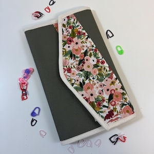 Pode incluir: Um estojo retangular de tecido verde-oliva com uma aba diagonal apresentando um fundo branco estampado com flores rosas, vermelhas e p&ecirc;ssego e folhas verdes. O estojo &eacute; debruado em tecido creme e cercado por pequenos marcadores de pontos.