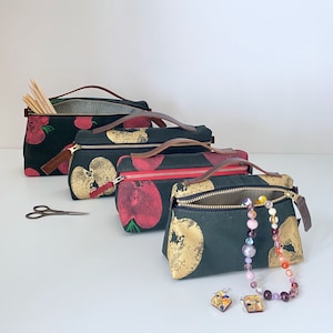 May include: Four black canvas zippered pouches with different patterns of red and gold apples. The pouches are lined up in a row from largest to smallest. The smallest pouch has a gold chain with colorful beads.