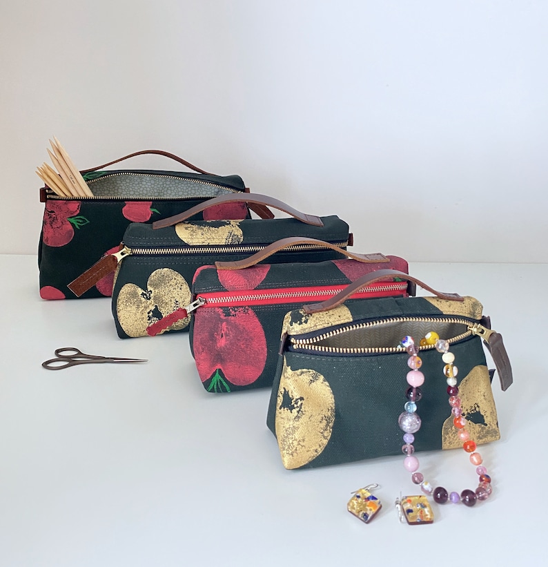 May include: Four black canvas zippered pouches with different patterns of red and gold apples. The pouches are lined up in a row from largest to smallest. The smallest pouch has a gold chain with colorful beads.