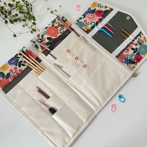 Pode incluir: Um estojo de tecido para organizar agulhas de tric&ocirc; e croch&ecirc; com um padr&atilde;o floral de flores rosas, azuis e amarelas sobre um fundo creme. O interior cont&eacute;m v&aacute;rios bolsos com diversas agulhas e ganchos de metal e madeira.